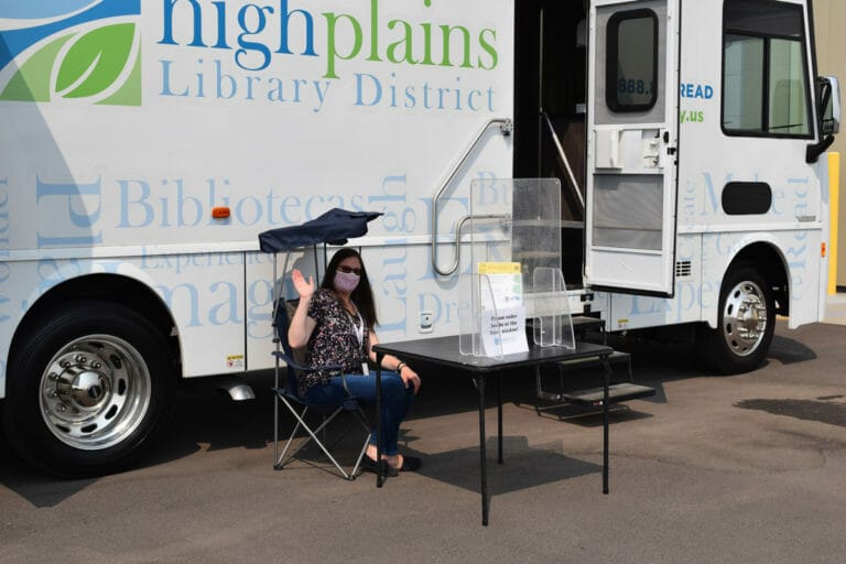 A woman waves in front of the High Plains Library District custom bookmobile.