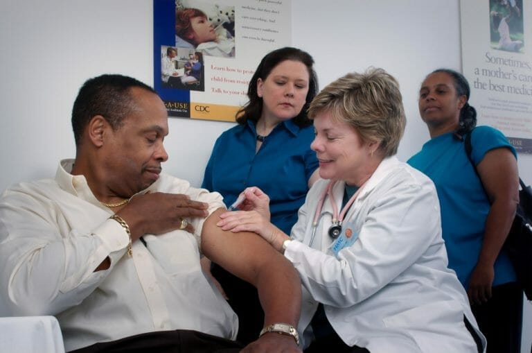 Female nurse or doctor giving a man a vaccination while two other medical professionals look on
